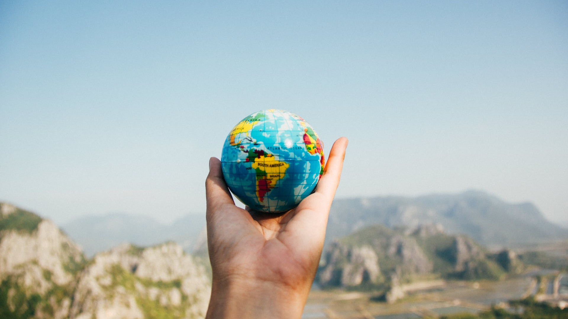 A hand holds up a toy globe in front of a mountainous background